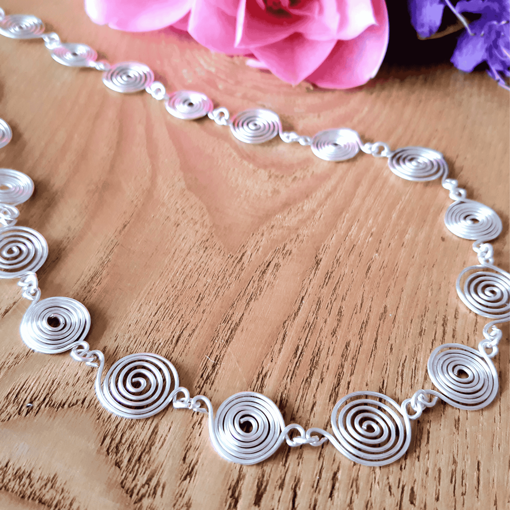 Silver spiral necklace on a wooden surface with pink flowers in the background