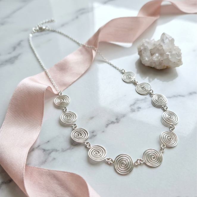 Silver necklace with circular design on a marble surface with pink ribbon and crystal.