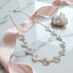 Silver necklace with circular design on a marble surface with pink ribbon and crystal.