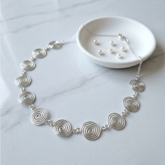 Silver spiral necklace on a light gray surface with a small white dish containing additional jewelry.