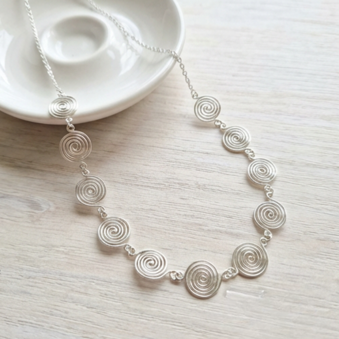 Silver spiral necklace on a light wooden surface with a white dish in the background.