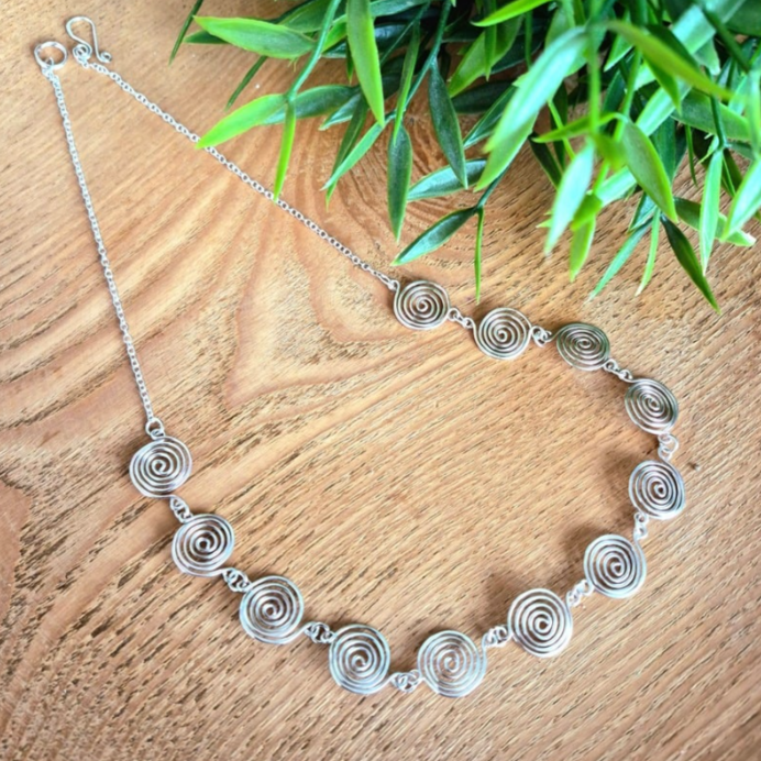 Silver necklace with circular patterns on a wooden surface with green leaves in the background