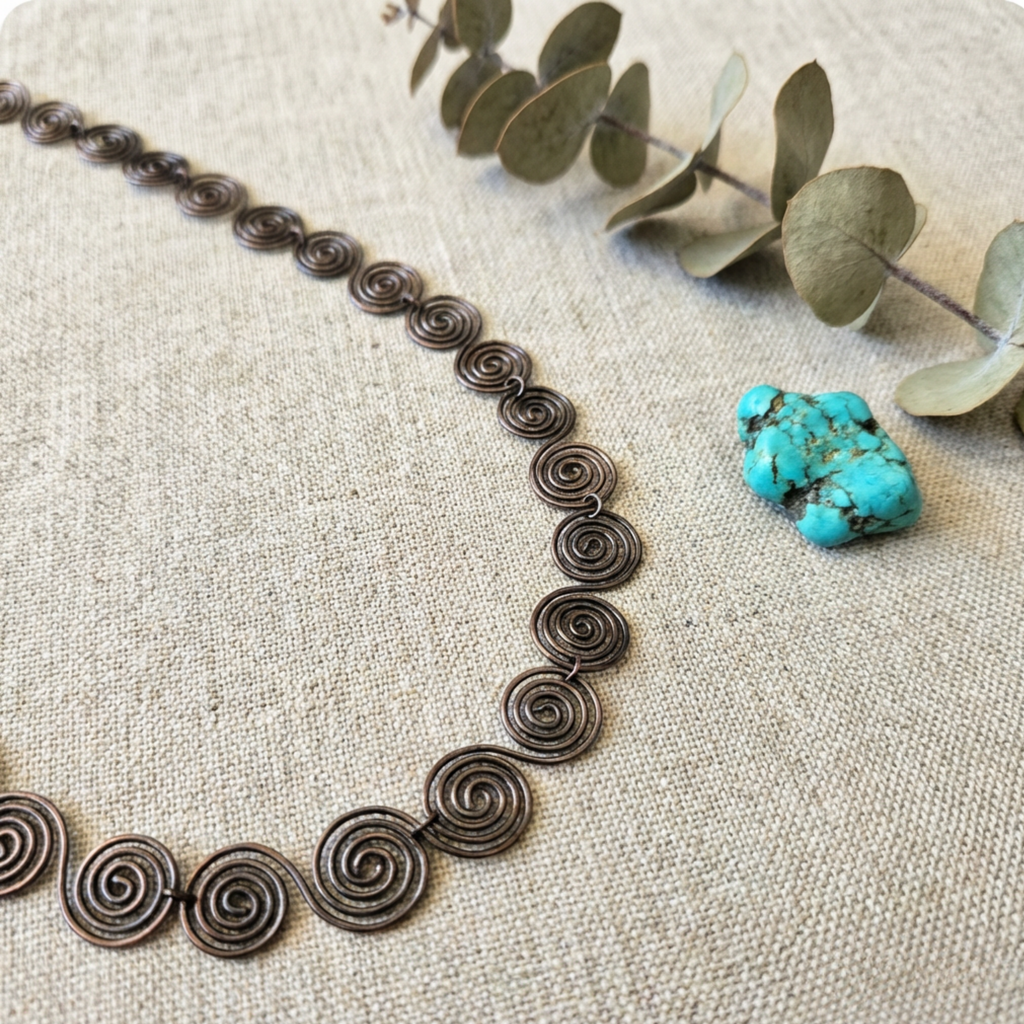 Necklace with spiral design on a textured surface with a turquoise stone and eucalyptus leaves.