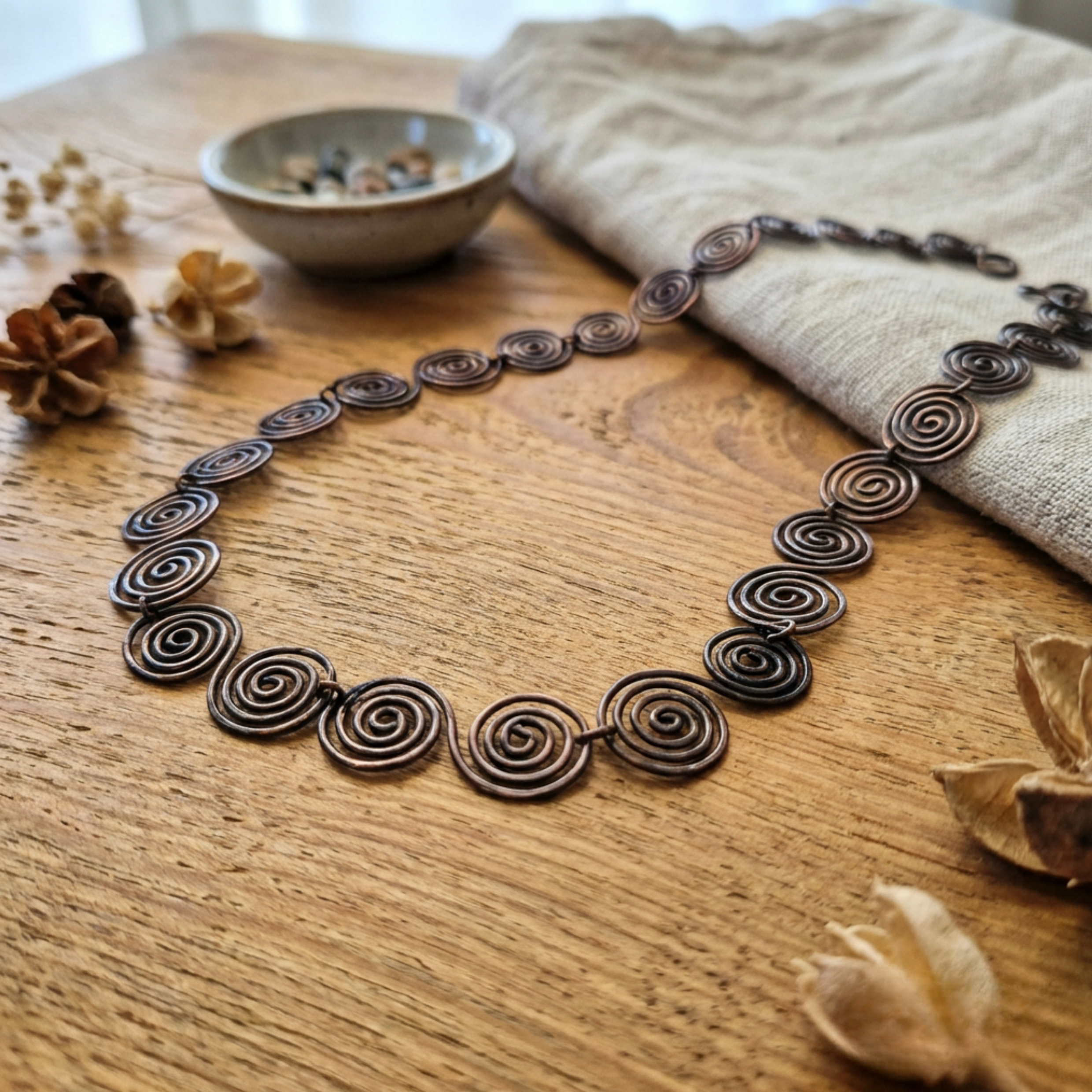 Copper necklace with spiral design on a wooden surface with dried flowers and a bowl.