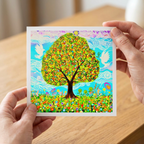 Colourful artwork of a tree and doves with abstract design greeting card held by hands on a wooden surface
