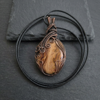 Brown picture jasper stone pendant with copper wire wrapping on a black cord against a dark stone background
