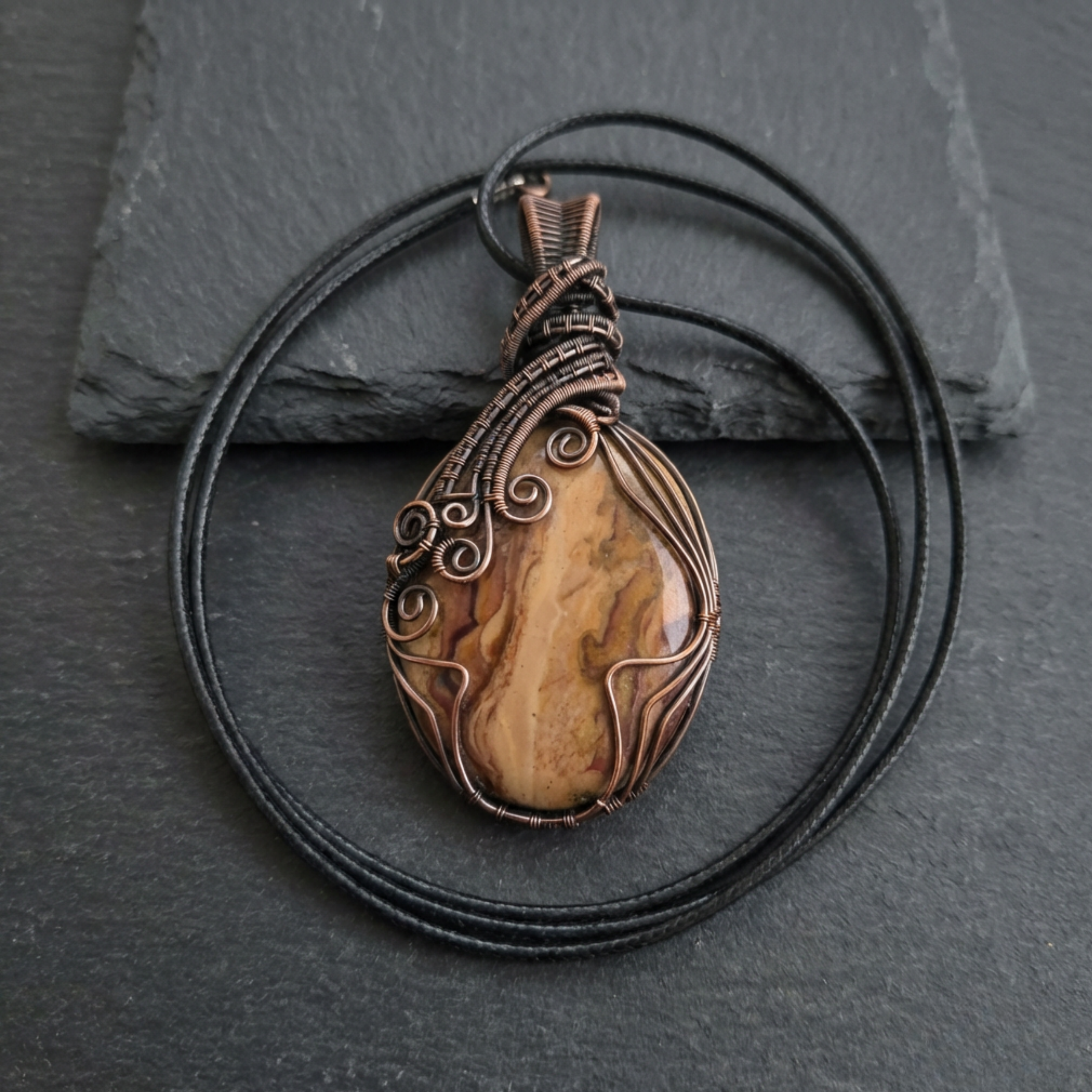 Brown picture jasper stone pendant with copper wire wrapping on a black cord against a dark stone background