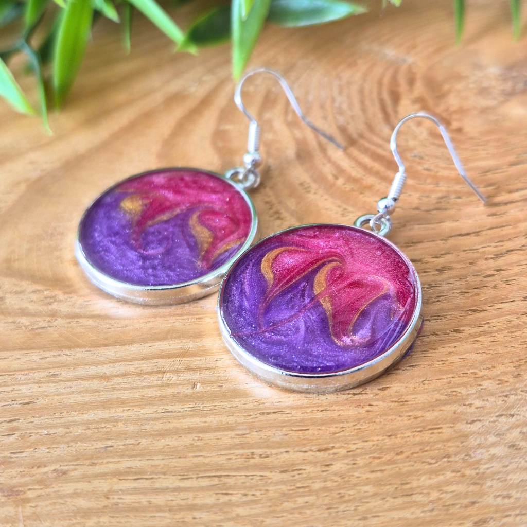 Purple and pink marbled earrings on a wooden surface with green leaves in the background