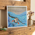 Greeting card artwork of a Puffin bird flying over water with colourful patterns on a wooden surface.