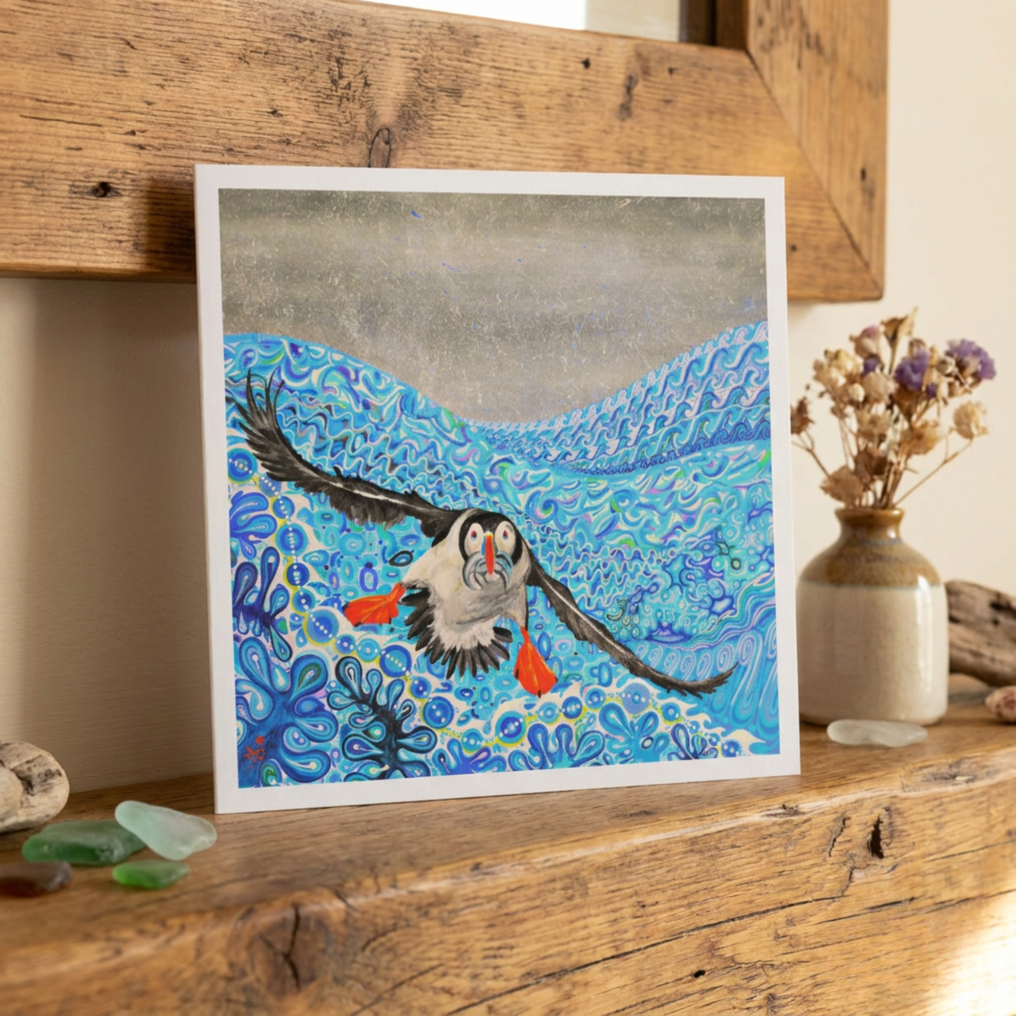 Greeting card artwork of a Puffin bird flying over water with colourful patterns on a wooden surface.