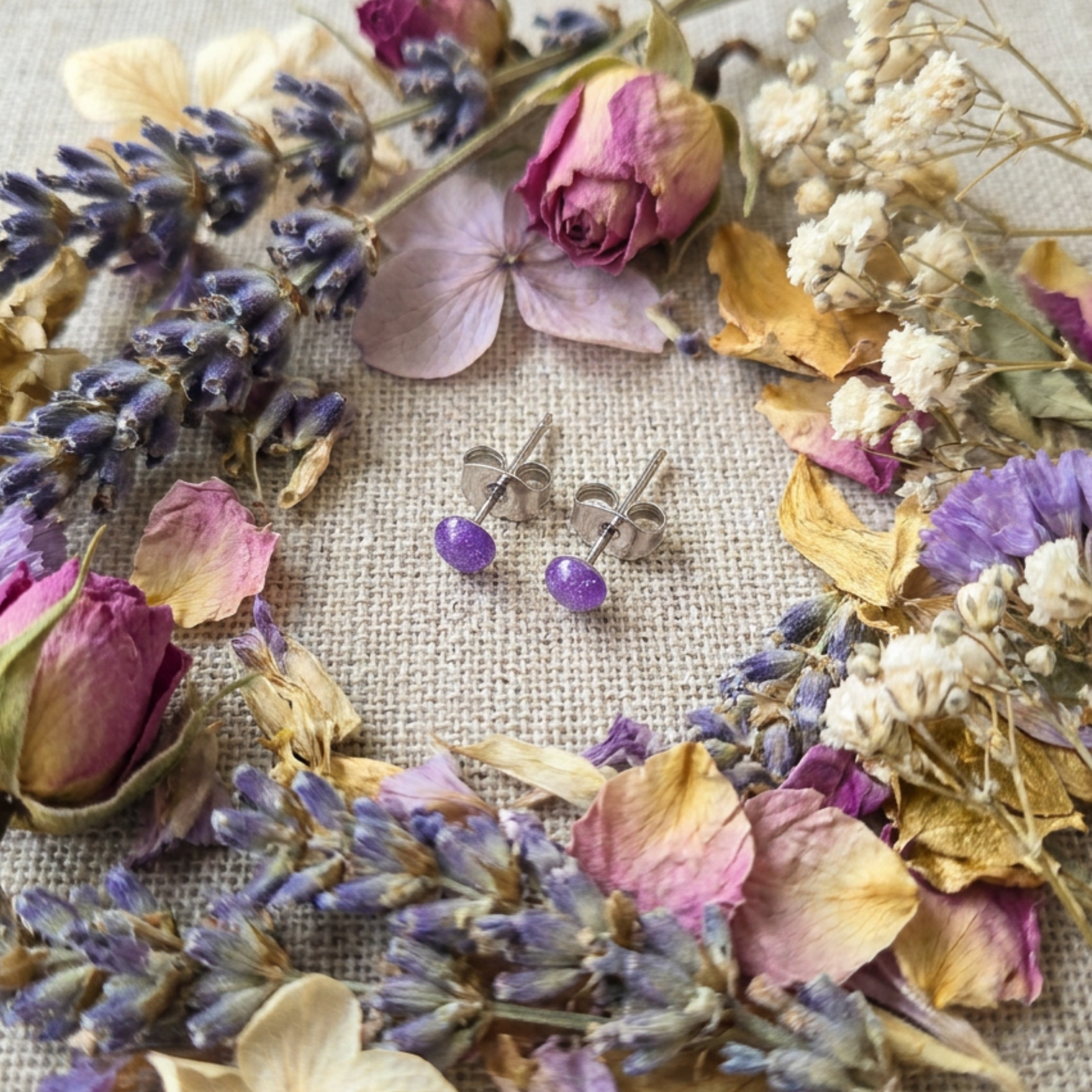 Silver earrings with purple stones surrounded by dried flowers on a textured fabric background