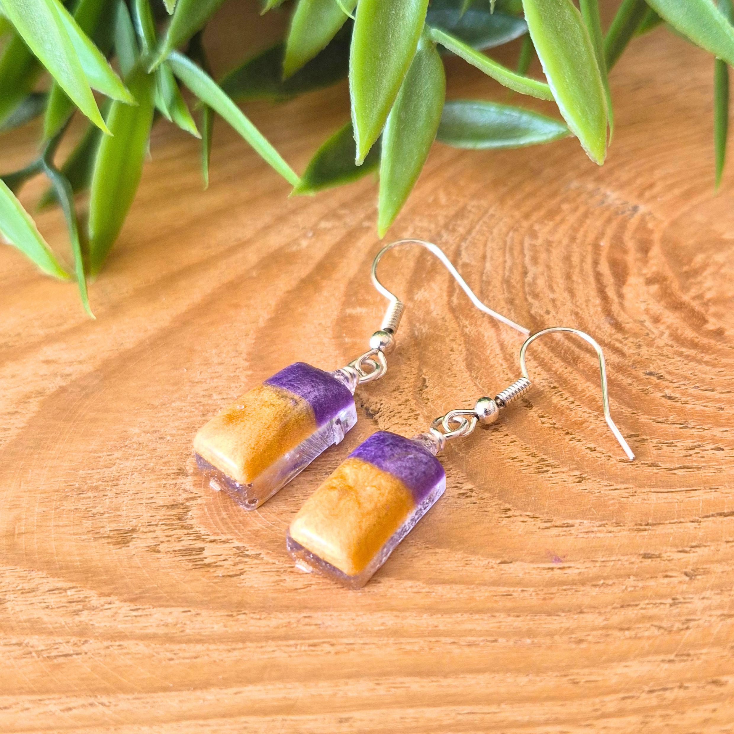 Pair of earrings with purple and gold rectangular stones on a wooden surface with green leaves in the background.