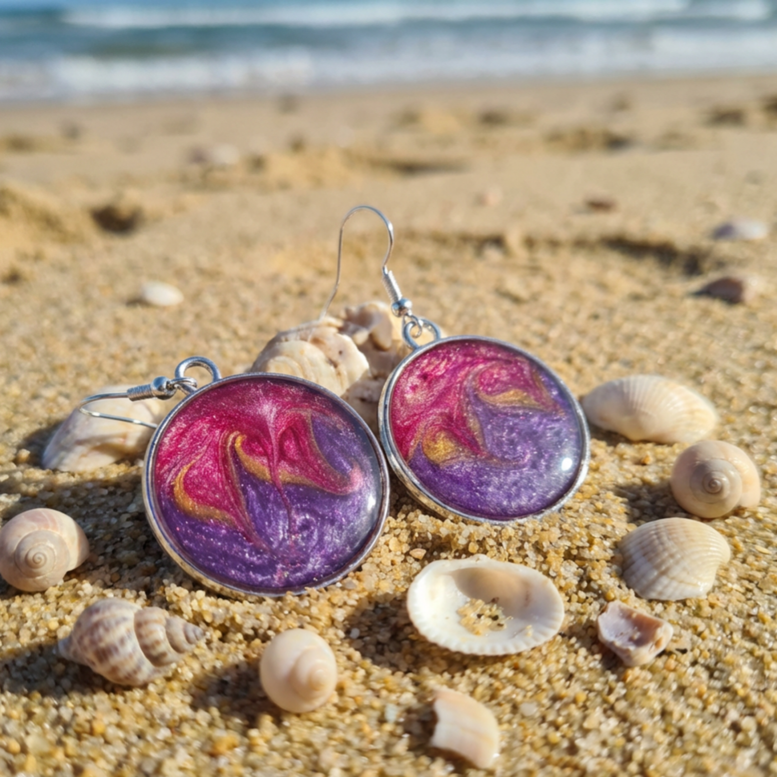 Purple and pink earrings on a sandy beach with shells