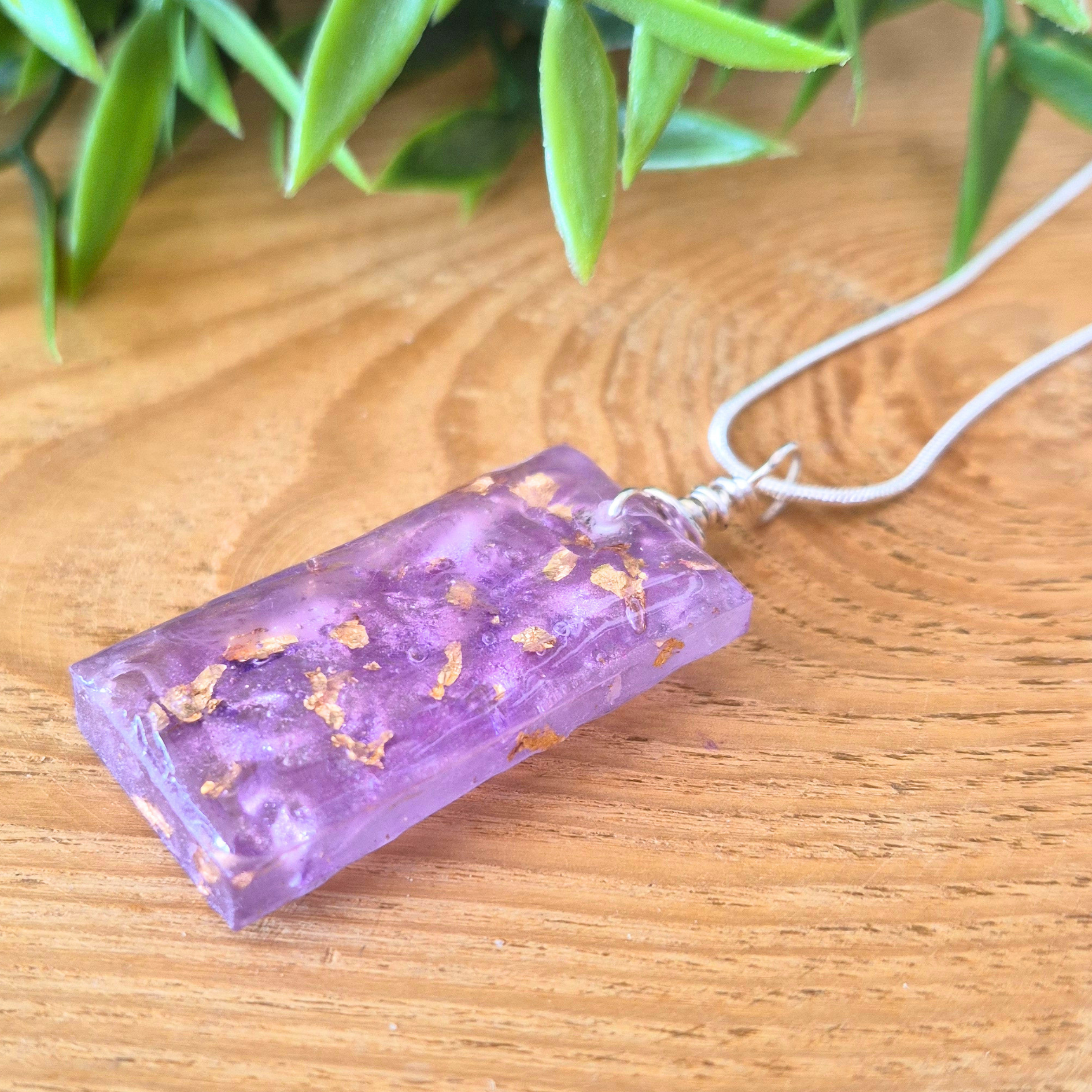 Purple rectangular pendant with gold flecks on a wooden surface with green leaves in the background