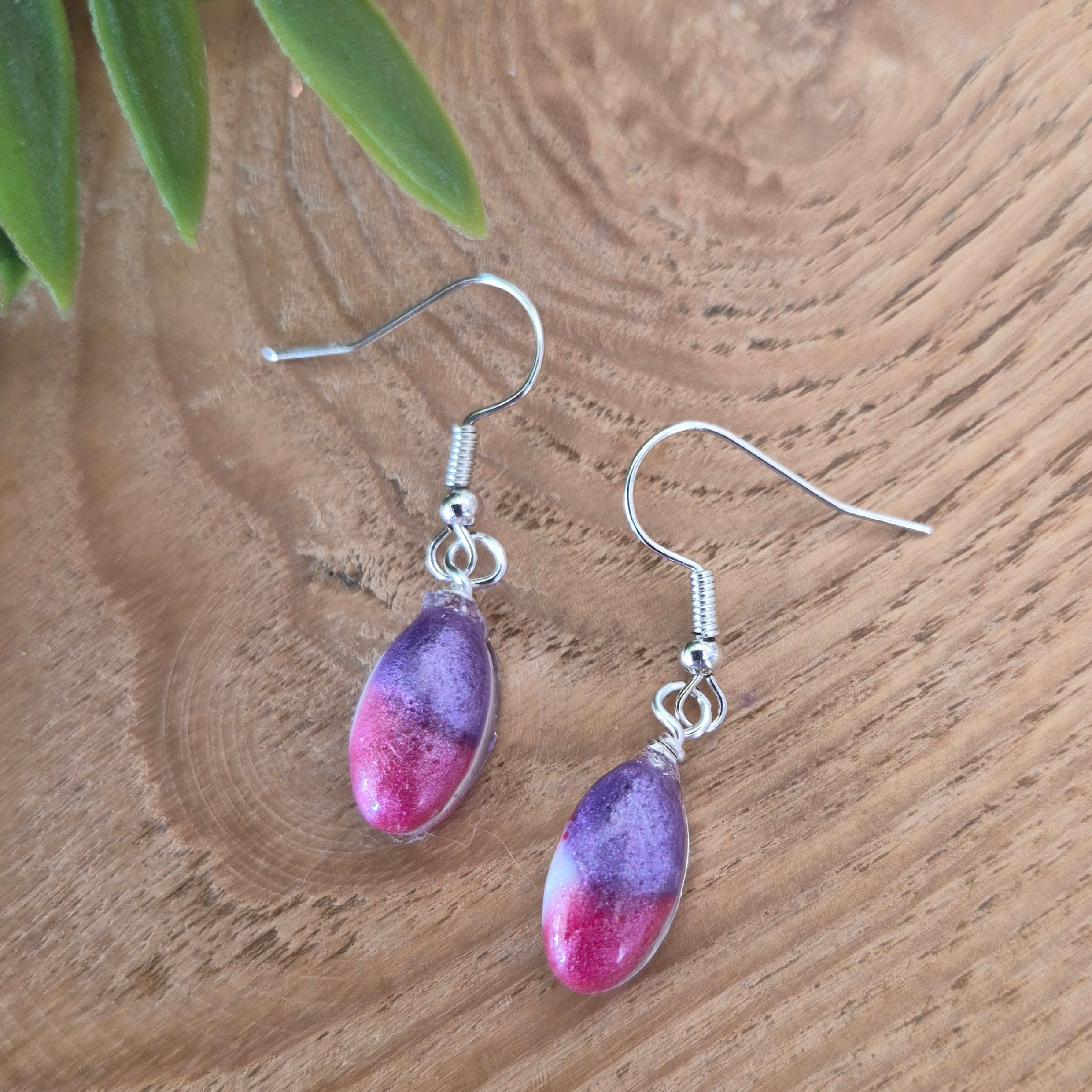 Purple and pink teardrop earrings on a wooden surface with green leaves.