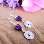 Purple heart-shaped earrings with silver spiral drops on a wooden surface with pink flowers.