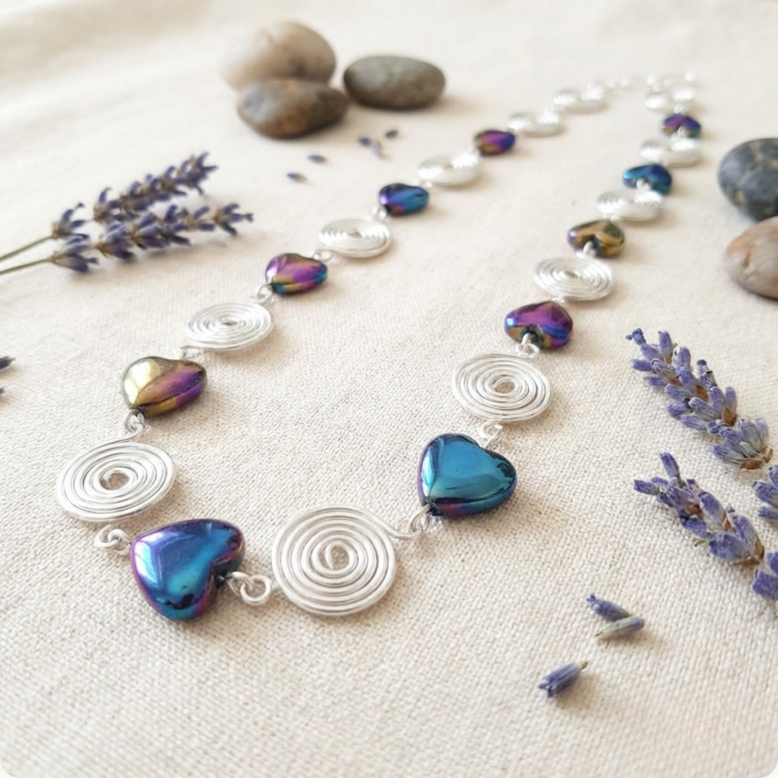 Necklace with heart-shaped beads and silver spirals on a textured surface with lavender flowers and stones