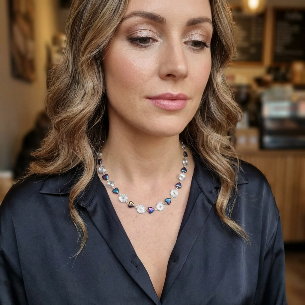 Woman wearing a silver spiral and iridescent heart bead necklace with a blurred indoor background