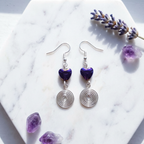 Pair of earrings with purple hematite heart stones and silver spiral designs on a marble surface with amethyst crystals.
