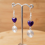 Pair of earrings with purple heart-shaped beads and silver spiral designs on a stand against a wooden background.