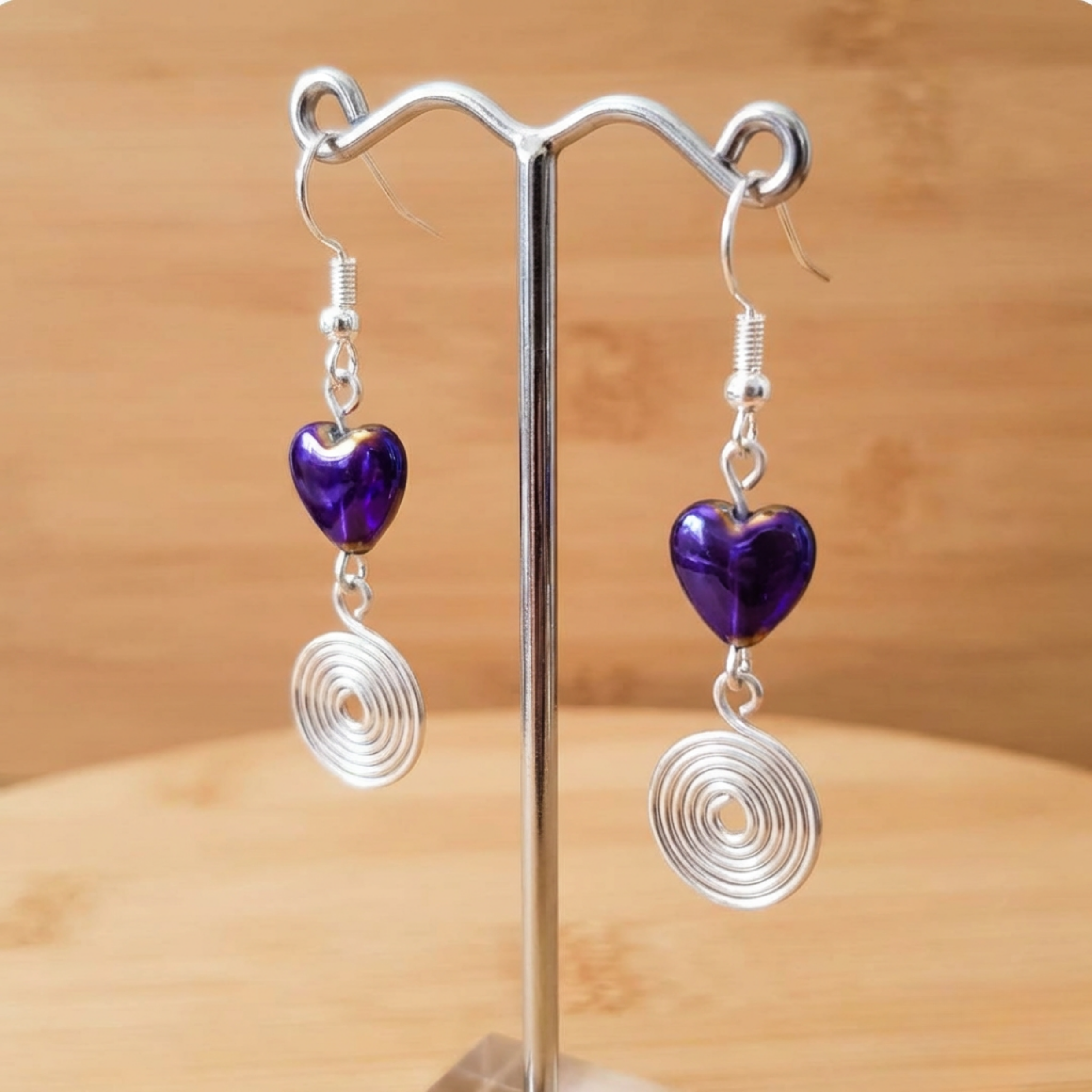 Pair of earrings with purple heart-shaped beads and silver spiral designs on a stand against a wooden background.