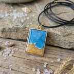 Gold Bezel seascape wave pendant on a black cord on stone and wood surface