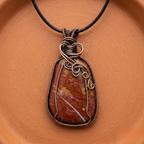 Jewellery pendant with brown red jasper stone and copper wire wrapping on a terracotta surface
