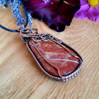 Red stone pendant with copper wire wrapping on a wooden surface with flowers in the background