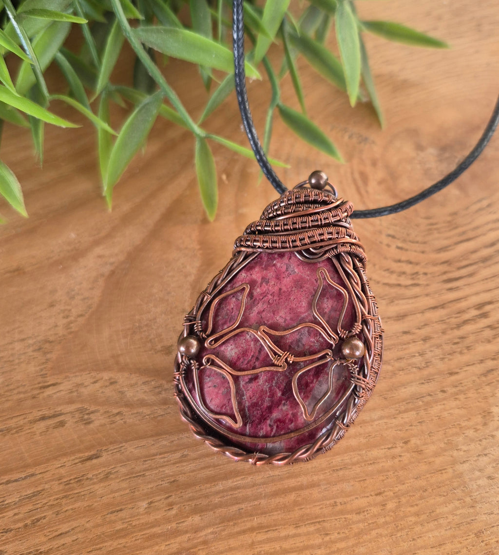 Pink stone pendant with copper wire wrapping on a wooden surface with green leaves in the background
