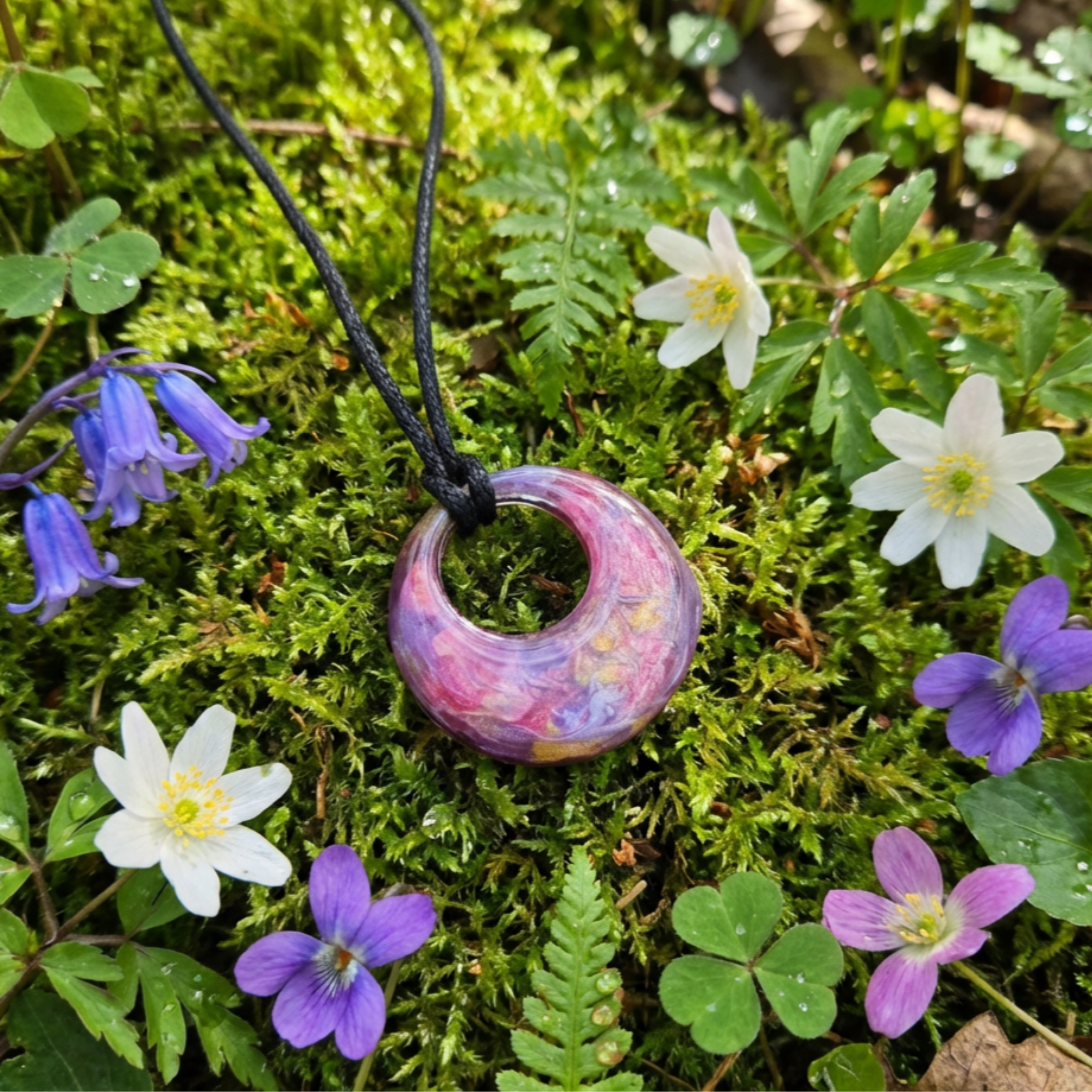 Pink and purple pendant on a black cord surrounded by green foliage and flowers