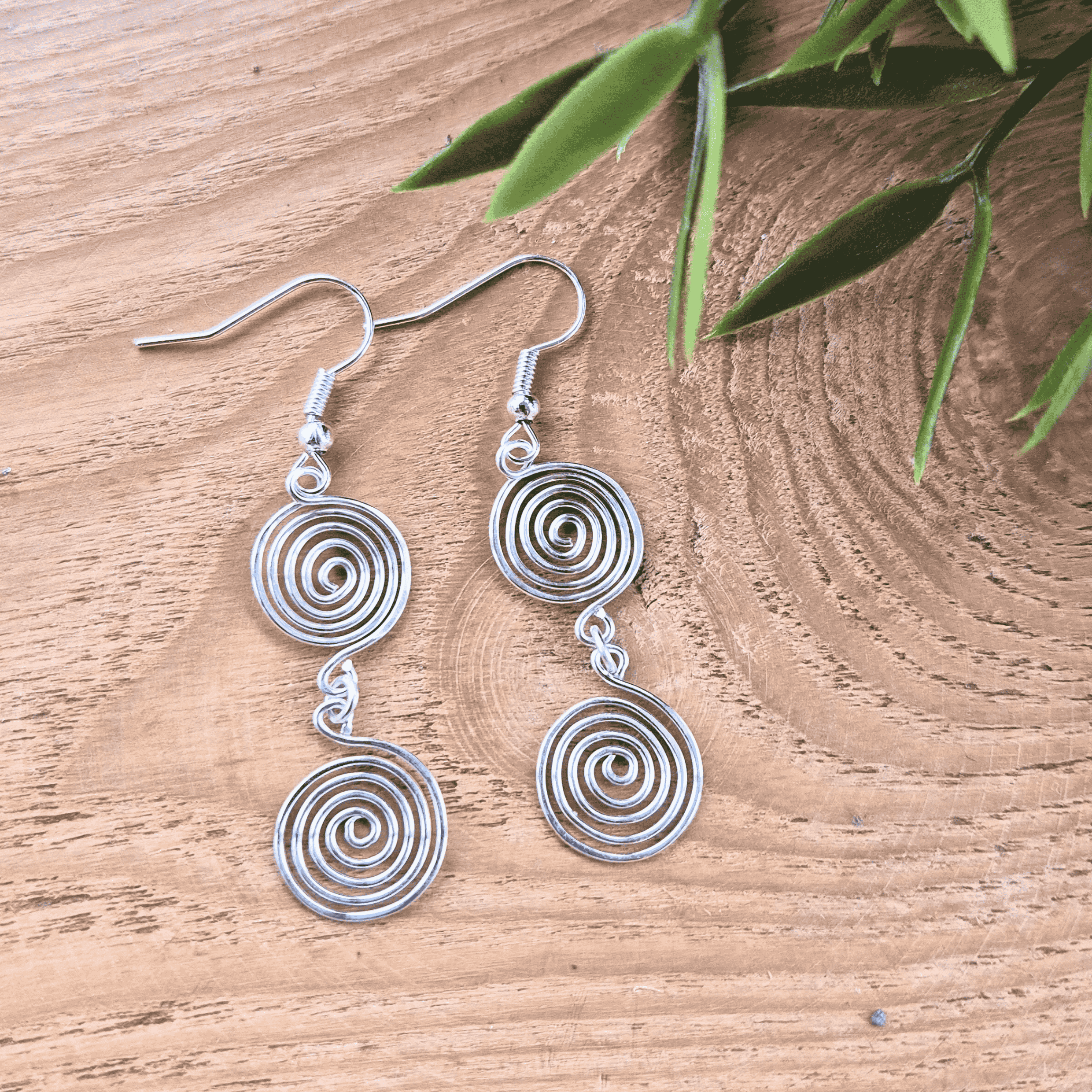 Silver spiral earrings on a wooden surface with green leaves.