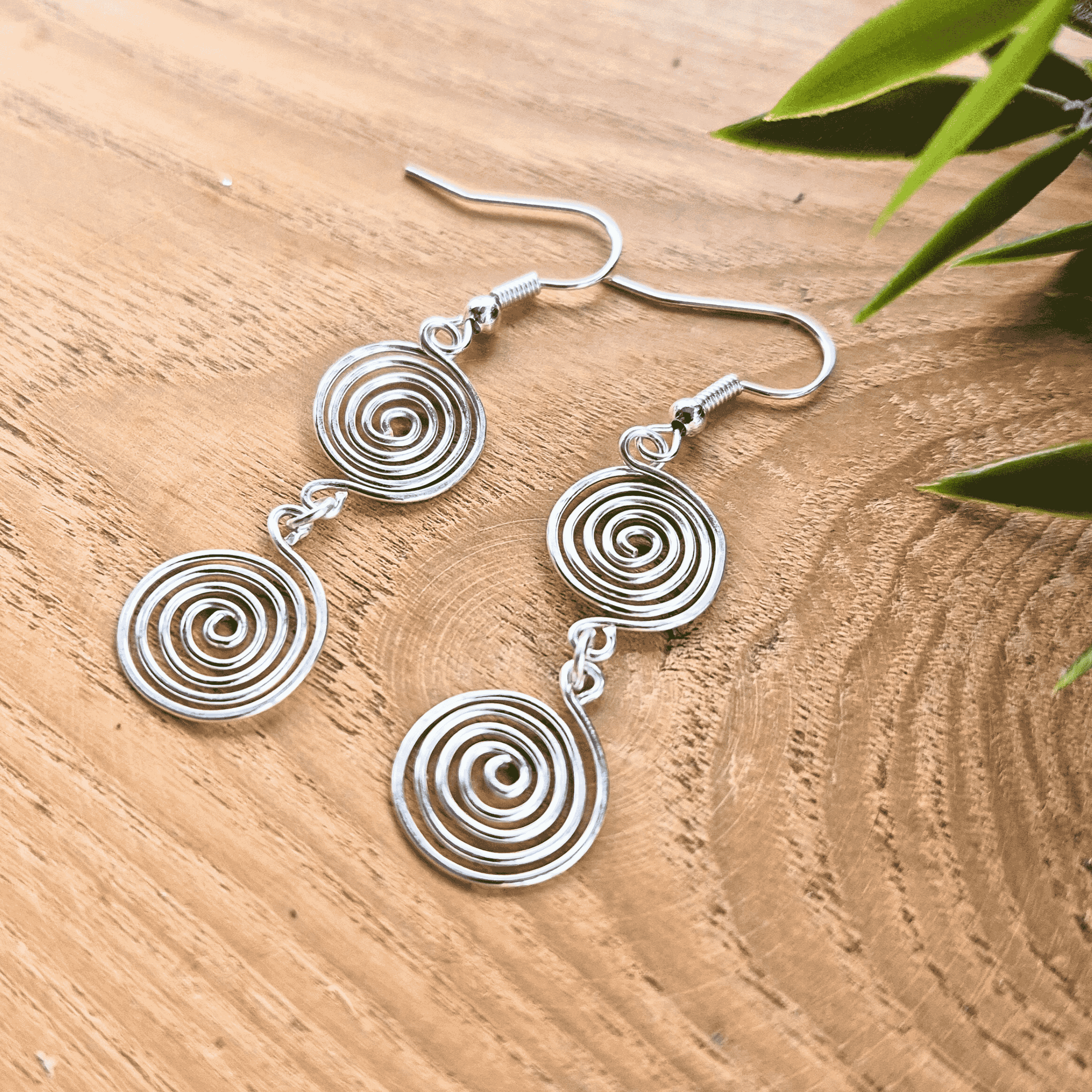 Silver spiral earrings on a wooden surface with green leaves.