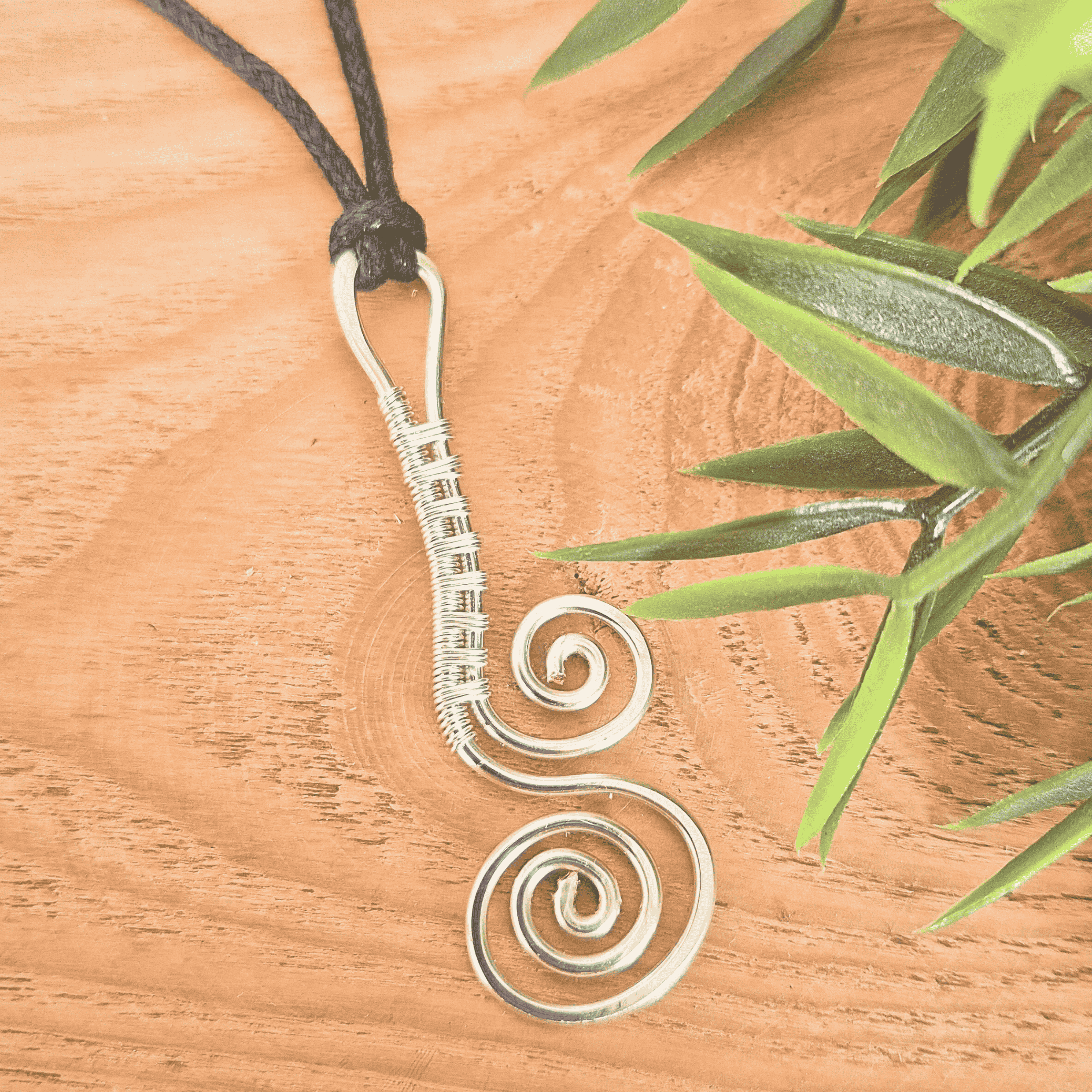 Silver spiral pendant on a black cord with green leaves on a wooden surface