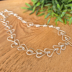 Silver necklace with heart designs on a wooden surface with green leaves in the background