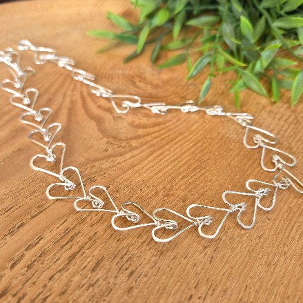Silver necklace with heart designs on a wooden surface with green leaves in the background