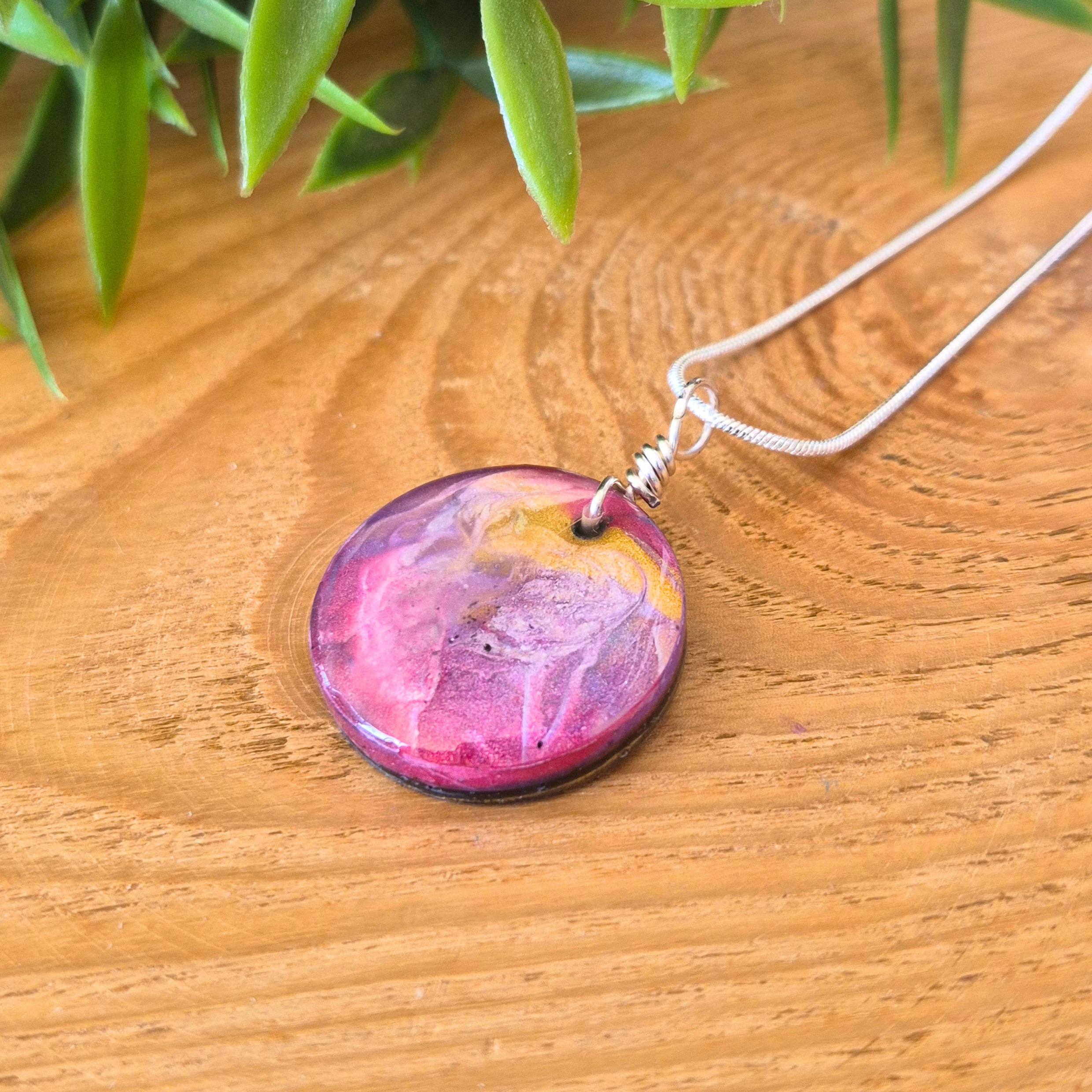 A small round resin pendant with pinks purples and gold spiralling on a wooden surface
