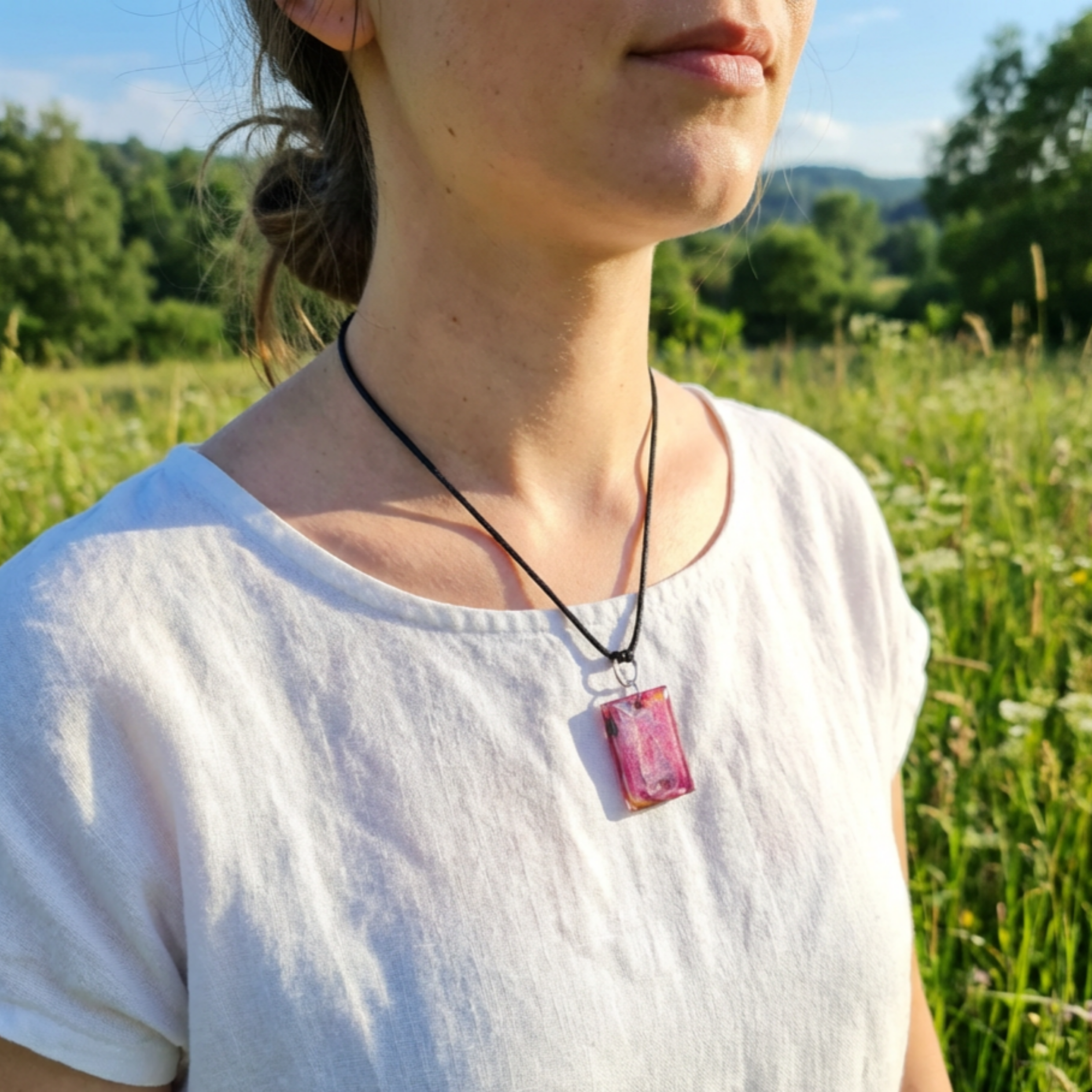 Lady wearing a pink rectangle pendant on a black cord standing in a meadow