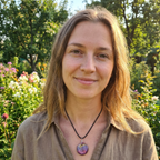 Woman with a necklace standing in a garden with flowers and trees.