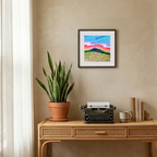 Wooden desk with a potted plant, typewriter, and framed artwork on a beige wall.