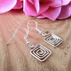 Silver spiral square earrings on a wooden surface with pink flowers in the background