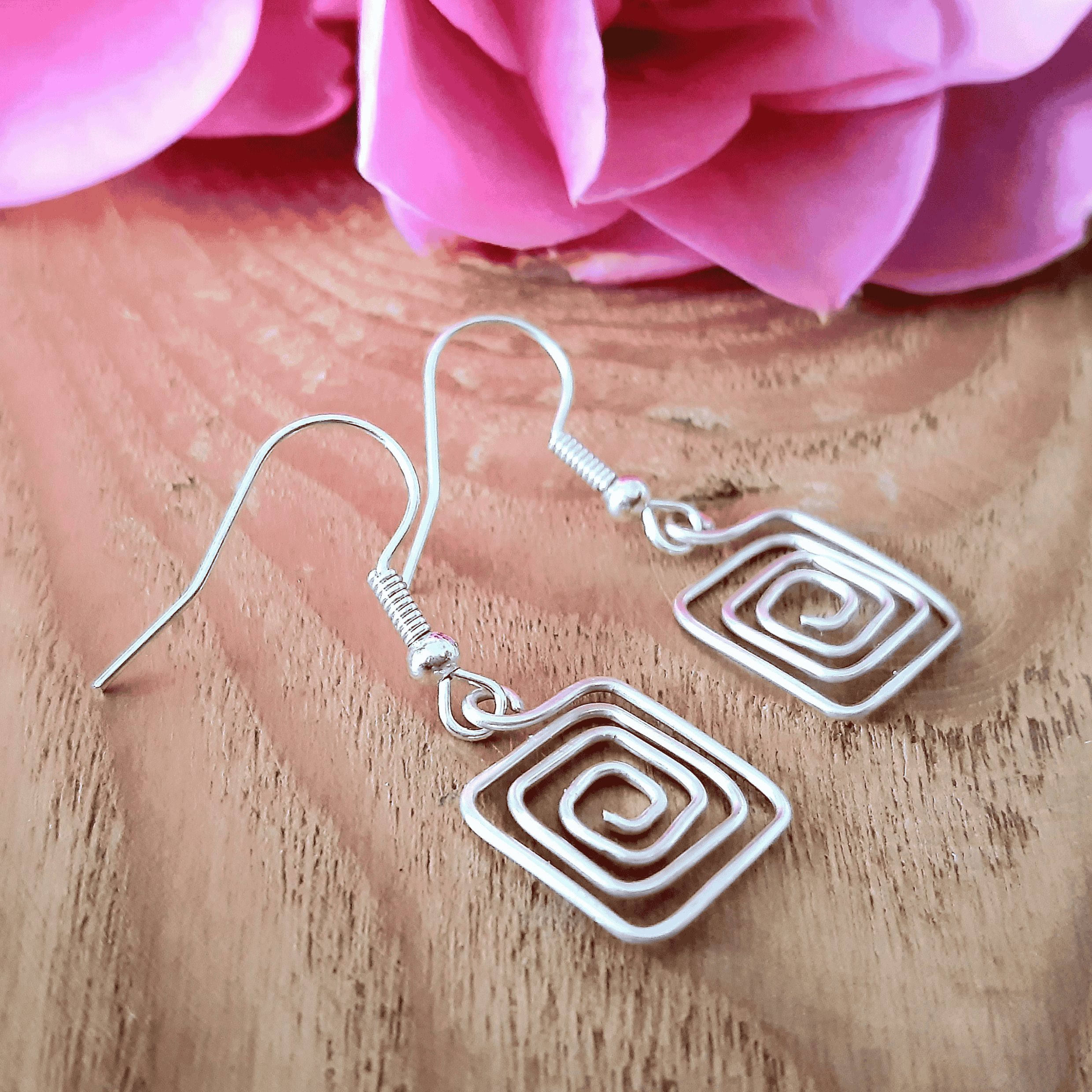 Silver spiral square earrings on a wooden surface with pink flowers in the background