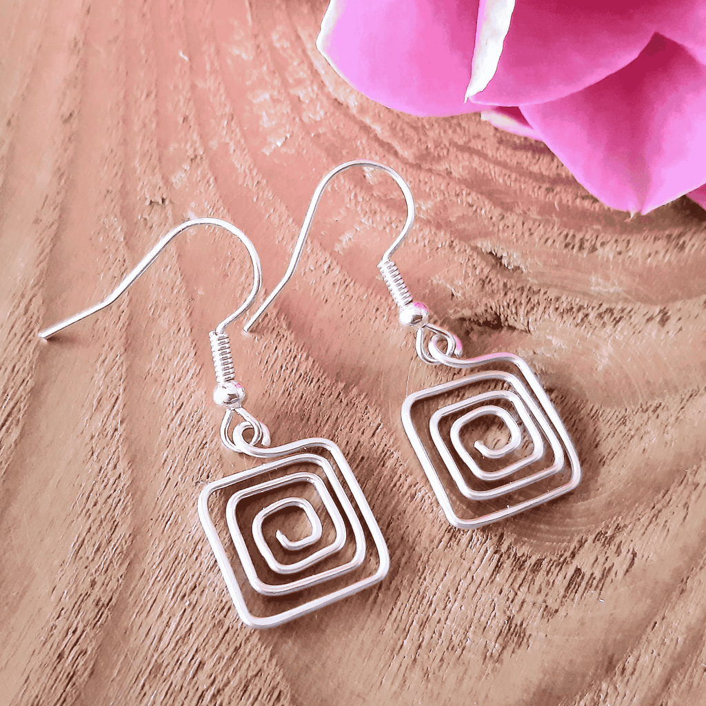 Silver spiral square earrings on a wooden surface with a pink flower.