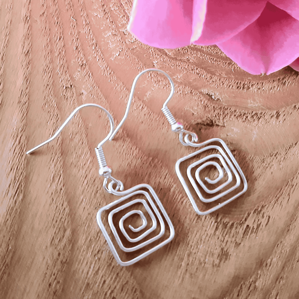 Silver square spiral earrings on a wooden surface with pink flowers.