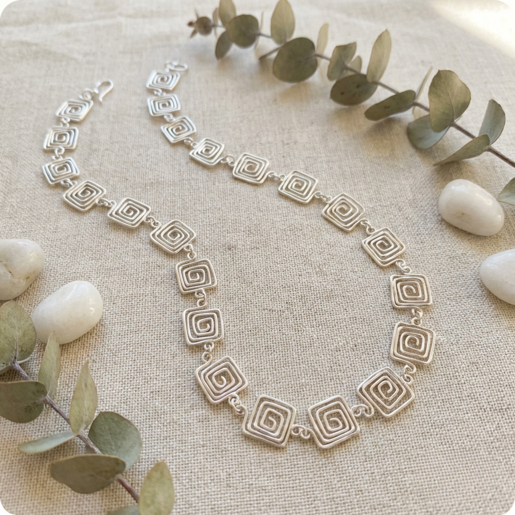 Silver necklace with geometric design on a textured surface with eucalyptus leaves and stones.