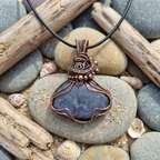 Copper wire-wrapped pendant with a dark stone on pebbles and driftwood