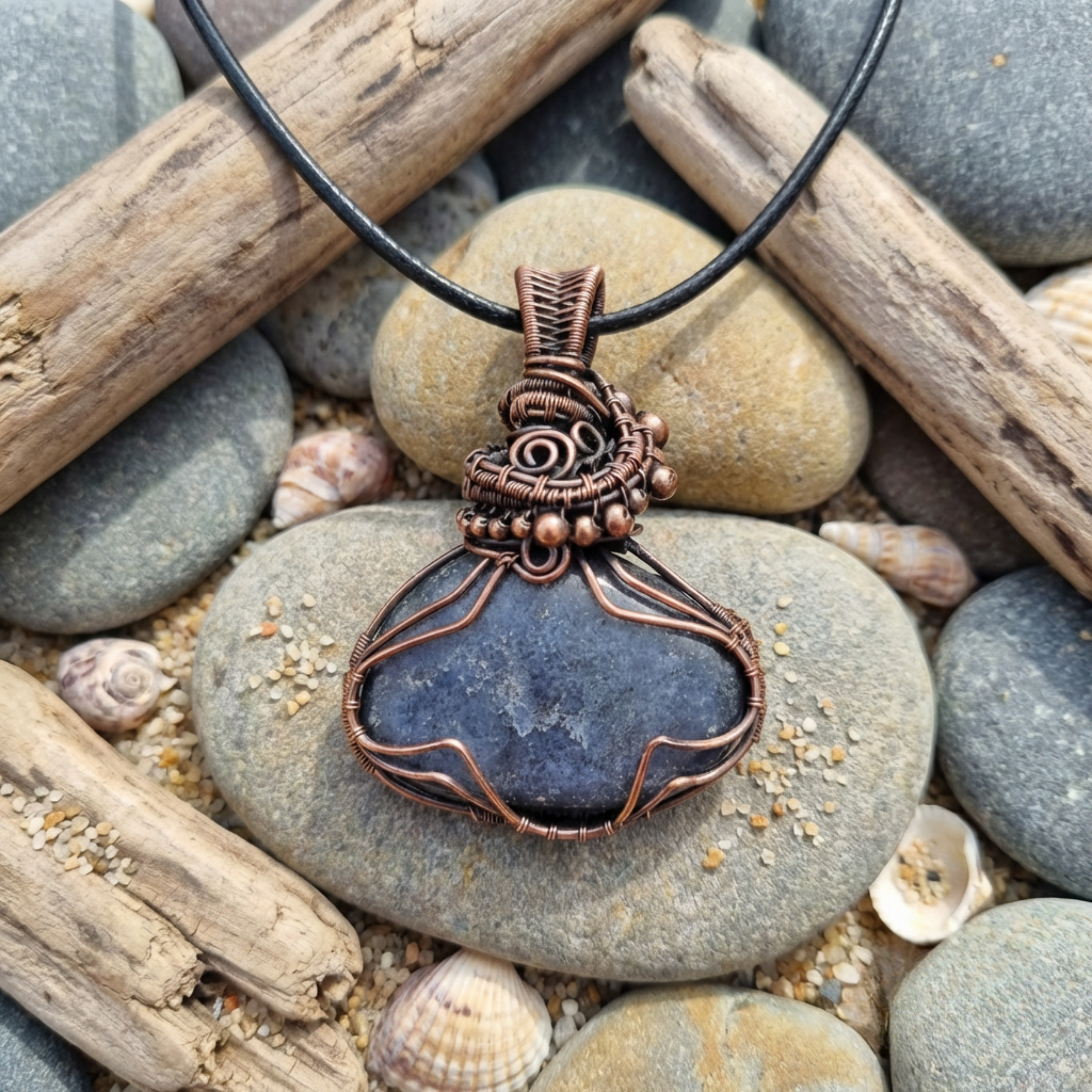 Copper wire-wrapped pendant with a dark stone on pebbles and driftwood