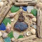 Pendant with blue stone and copper wirework on a sandy background with shells and driftwood