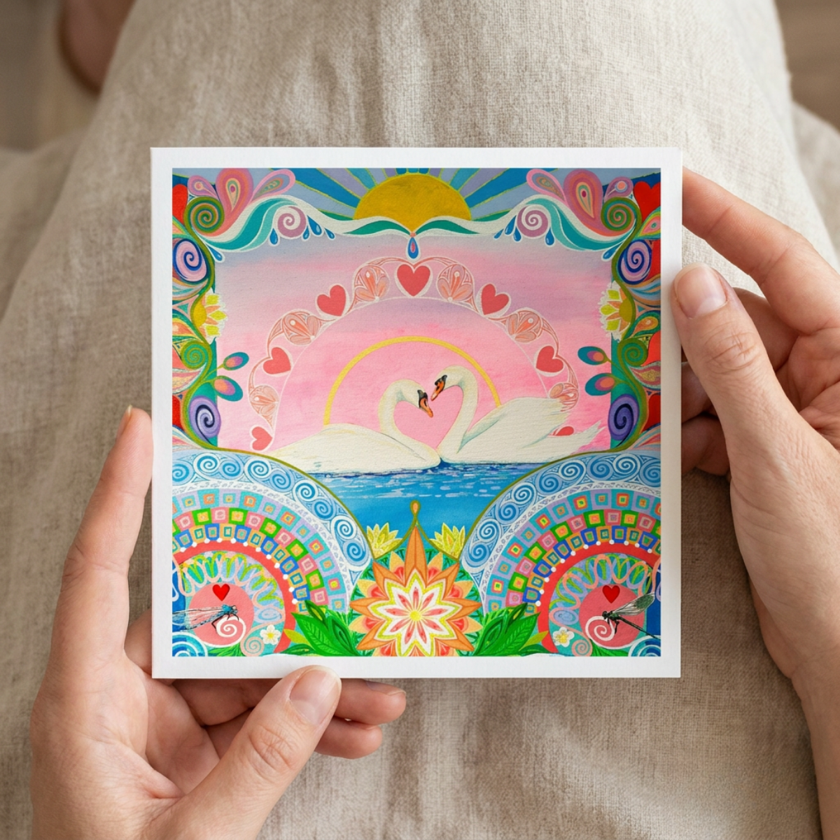 Colourful artwork of swans and abstract patterns greeting card held by hands against a neutral background