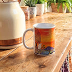 Colourful mug with a scenic design on a wooden table next to a white kettle.