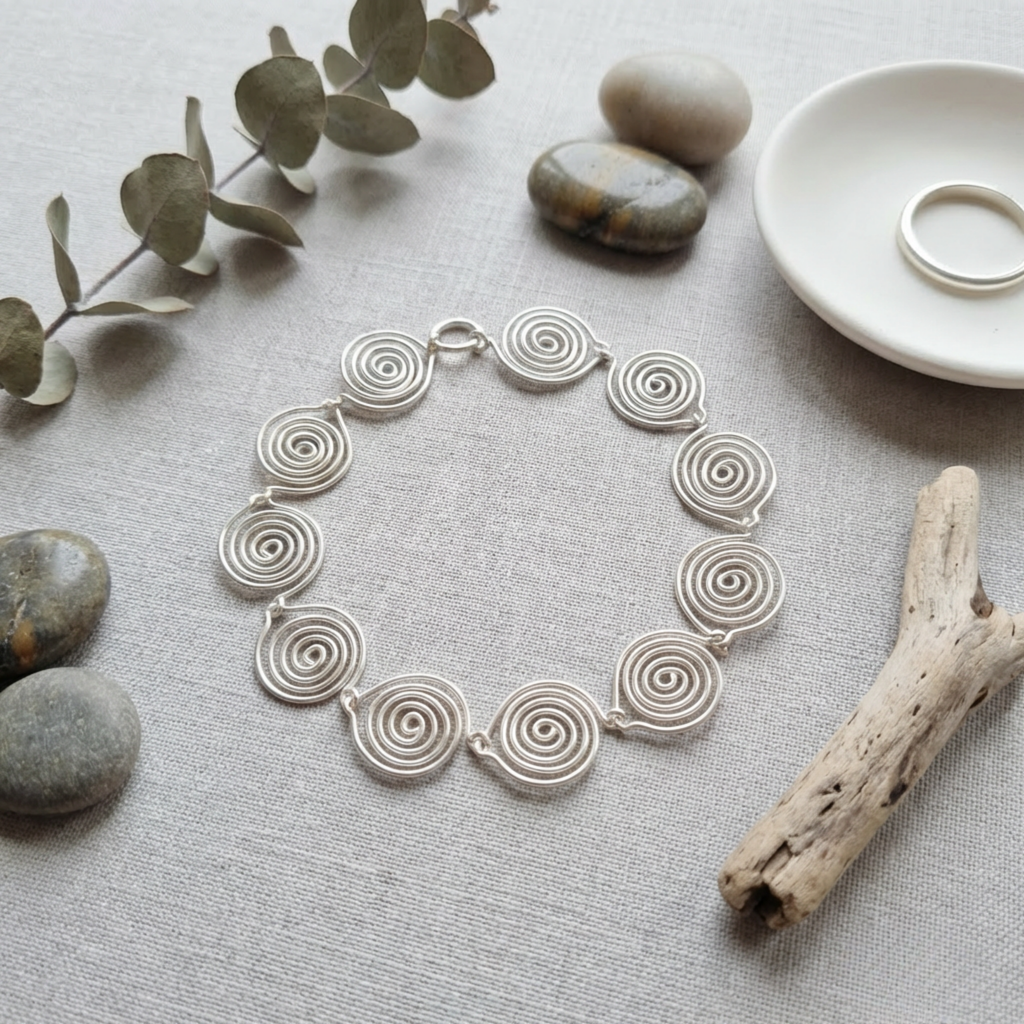 Silver spiral necklace on a textured surface with stones and a branch.